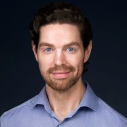 Roar has blue eyes, dark curly hair, and a beard. He is wearing a blue shirt.