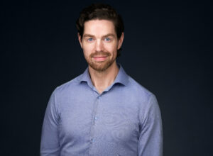 Roar has blue eyes, dark curly hair, and a beard. He is wearing a blue shirt.